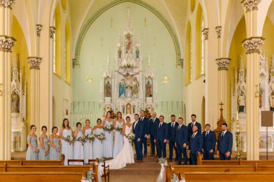 A wedding party poses inside a church with high arches and a large ornate altar. The bridesmaids wear light blue dresses, and the groomsmen wear navy suits. The bride and groom stand together at the center. by Justin Salem Meyer