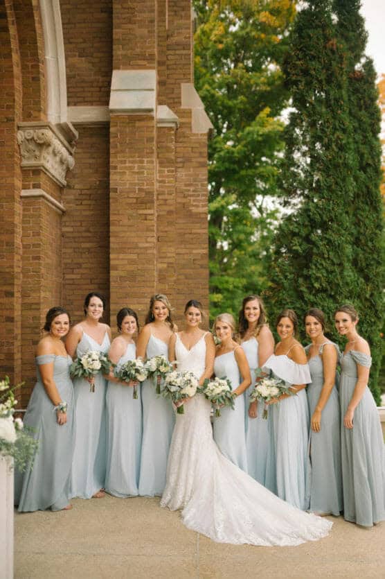 A bride in a white gown stands with nine bridesmaids in pale blue dresses, all holding bouquets, posing together outside a brick building with greenery in the background. by Justin Salem Meyer