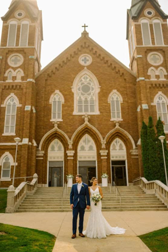 A bride and groom stand hand in hand outside a large brick church with arched doorways and tall twin towers, dressed in wedding attire on a sunny day. by Justin Salem Meyer