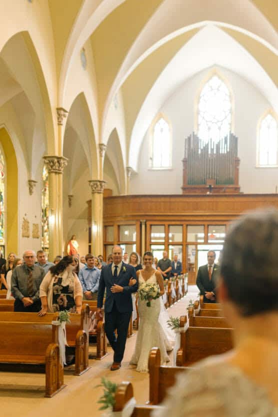 A bride in a white gown walks down the aisle of a church, holding a bouquet and escorted by a man in a blue suit, as guests seated in pews look on. Sunlight streams through tall windows, illuminating the space. by Justin Salem Meyer