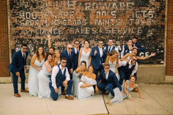 A joyful wedding party poses playfully in front of a vintage brick wall. The group is dressed in light blue dresses and navy suits, some wearing sunglasses, as they smile, laugh, and strike fun poses on a sidewalk. by Justin Salem Meyer