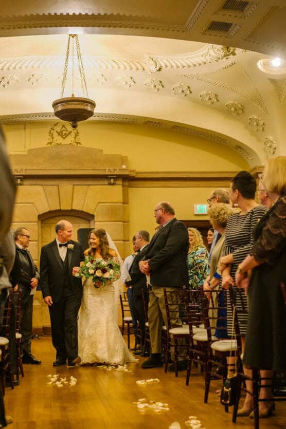 A bride in a white gown and veil walks down the aisle with an older man in a tuxedo, surrounded by seated and standing guests in a decorated indoor venue with ornate ceilings. by Justin Salem Meyer