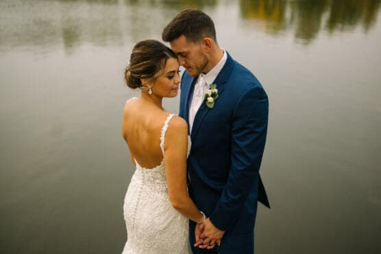 A bride in a white lace dress and a groom in a blue suit stand close together by a calm lake, gently holding hands and touching foreheads, sharing an intimate moment. by Justin Salem Meyer