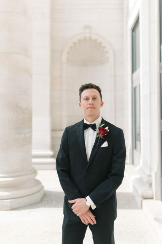 A man in a black tuxedo with a white shirt and black bow tie stands in a bright, elegant hallway with tall columns. He has a dark red and white boutonniere pinned to his lapel. by Justin Salem Meyer