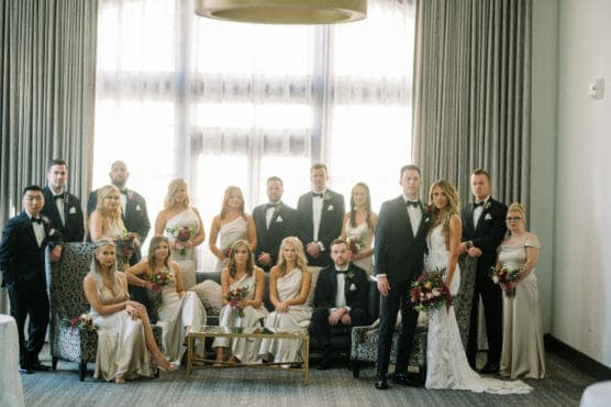 A wedding party poses indoors in front of a large window. The group includes bridesmaids in light dresses holding bouquets and groomsmen in black tuxedos. The bride and groom stand in the center, with the group arranged around them. by Justin Salem Meyer