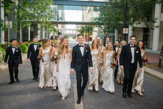 A bride and groom, dressed in wedding attire, walk joyfully down a city street with their wedding party. Bridesmaids wear matching light dresses, and groomsmen wear black tuxedos. The group smiles and laughs together outdoors. by Justin Salem Meyer