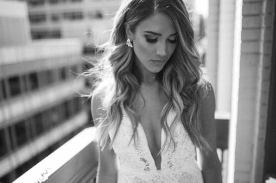A woman in a white lace dress with long wavy hair stands on a balcony, looking down with a serene expression. The image is in black and white, capturing a soft and elegant atmosphere. by Justin Salem Meyer