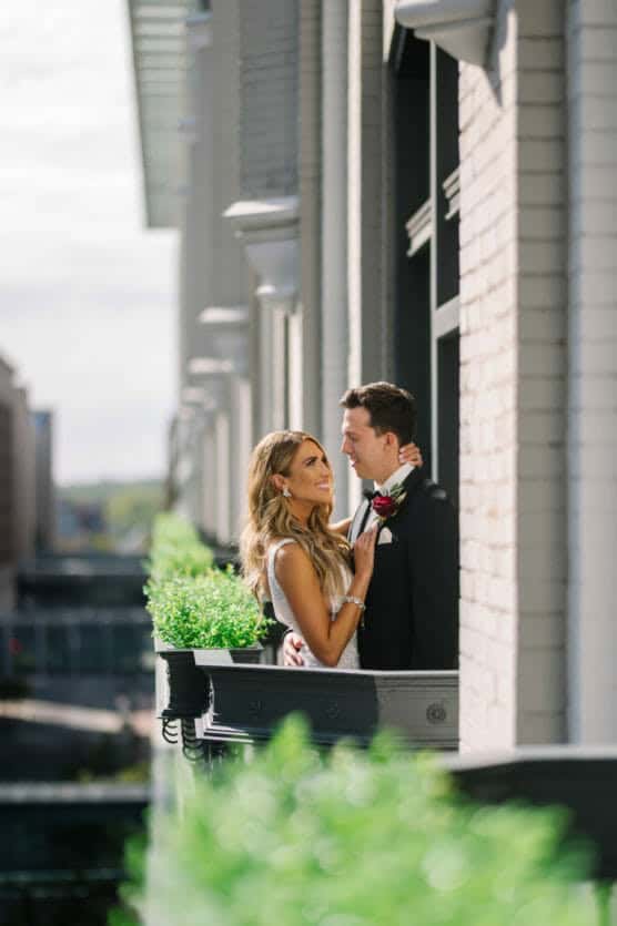 A bride and groom stand closely together on a small balcony, smiling at each other. The bride holds the groom’s lapel, and greenery lines the balcony railing. The background shows an urban cityscape. by Justin Salem Meyer
