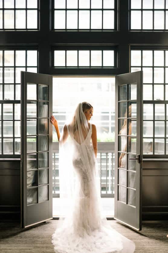 A bride in a white lace gown and veil stands in front of open double doors, looking outside. Sunlight streams in through tall grid windows, creating a bright, elegant atmosphere. by Justin Salem Meyer