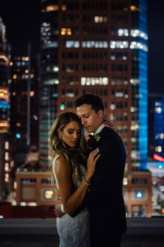 A bride and groom embrace tenderly on a city rooftop at night, with tall, illuminated buildings in the background creating a romantic urban atmosphere. by Justin Salem Meyer