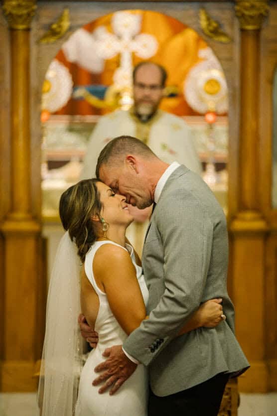 A bride and groom embrace and touch foreheads, smiling lovingly, during their wedding ceremony in a church, with an officiant and ornate altar in the background. by Justin Salem Meyer