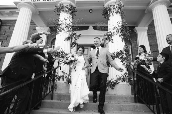 A newlywed couple walks down the steps outside a building as guests cheer and throw confetti. The bride holds a bouquet and laughs, while the groom smiles. The scene is joyful with greenery decorating the pillars. by Justin Salem Meyer