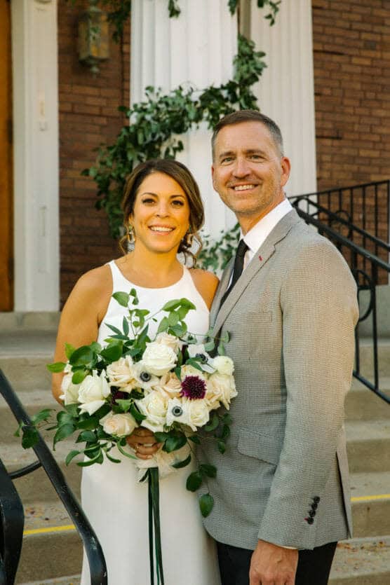 A bride in a white dress holding a bouquet of white flowers stands next to a groom in a light gray suit and black tie. They are smiling, posing outdoors in front of a brick building with white columns and greenery. by Justin Salem Meyer