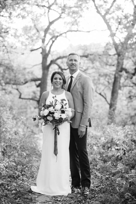A bride in a white dress holding a bouquet stands next to a groom in a suit and tie. They are outdoors on a wooded path with sunlight filtering through the trees. The photo is in black and white. by Justin Salem Meyer