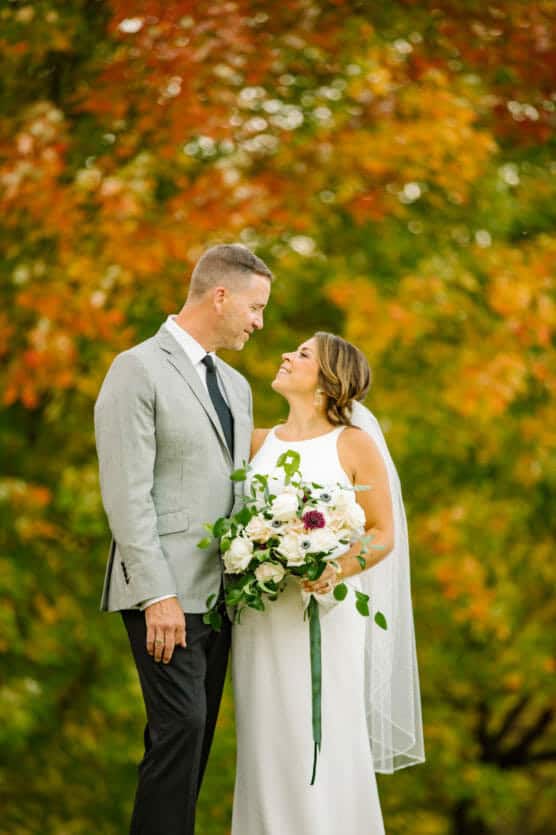 A bride and groom stand outdoors, smiling at each other, surrounded by vibrant autumn foliage. The bride holds a bouquet of white flowers and wears a white dress with a veil, while the groom wears a light gray jacket and black tie. by Justin Salem Meyer