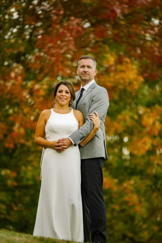 A couple stands outdoors in front of colorful autumn foliage. The woman wears a white sleeveless dress, and the man wears a light gray suit. They smile, standing close together and holding hands. by Justin Salem Meyer