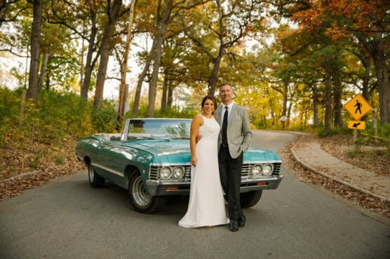 A couple dressed in formal attire stands in front of a vintage turquoise convertible on a winding road surrounded by autumn trees. The woman wears a white dress and the man wears a light gray jacket. by Justin Salem Meyer