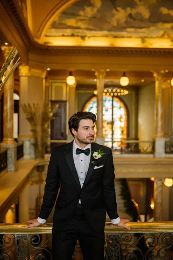 A man in a black tuxedo with a boutonniere stands in an ornate, golden hall with marble columns and a stained glass window in the background, looking to the side. by Justin Salem Meyer