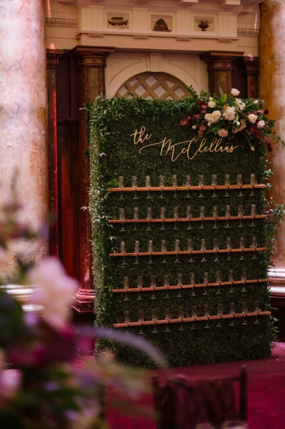 A decorated champagne wall with rows of filled glasses and a lush greenery backdrop is topped with flowers and a sign reading the McClellans, set in an elegant, ornate venue. by Justin Salem Meyer