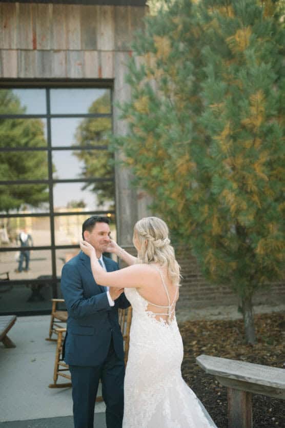 A bride in a lace wedding gown gently holds the face of a groom in a blue suit outside, near a tall pine tree and a building with large windows reflecting the scene. by Justin Salem Meyer