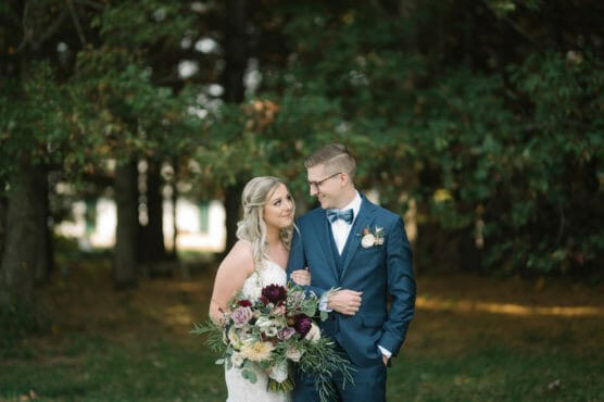 A bride in a white dress holding a bouquet of flowers stands arm-in-arm with a groom in a navy suit and bow tie. They smile at each other, surrounded by lush greenery and trees in the background. by Justin Salem Meyer