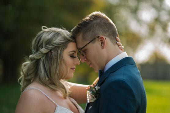 A bride and groom stand close together outdoors, touching foreheads and closing their eyes, sharing an intimate, tender moment on their wedding day. Soft sunlight and greenery are visible in the background. by Justin Salem Meyer