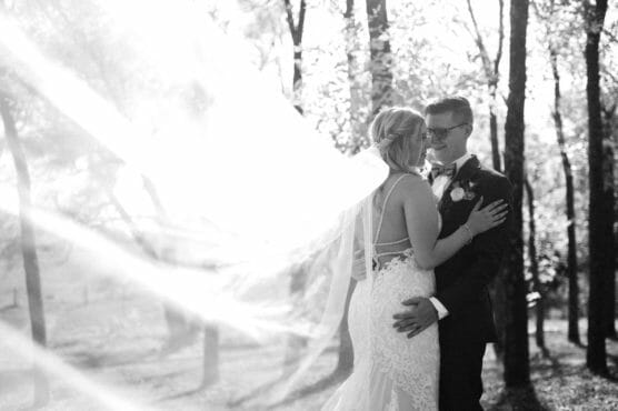 A bride and groom embrace outdoors among trees, smiling at each other. Sunlight shines through the bride’s veil, creating a soft, glowing effect. The scene is romantic and joyful. by Justin Salem Meyer