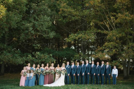 A wedding party poses outdoors in front of tall green trees. The bride and groom stand in the center, surrounded by bridesmaids in pink and blue dresses and groomsmen in blue suits. A young boy stands at the far right. by Justin Salem Meyer