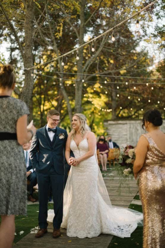 A bride in a white lace gown stands smiling beside the groom in a navy suit during an outdoor wedding ceremony, surrounded by trees and string lights, with guests and bridesmaids in the background. by Justin Salem Meyer