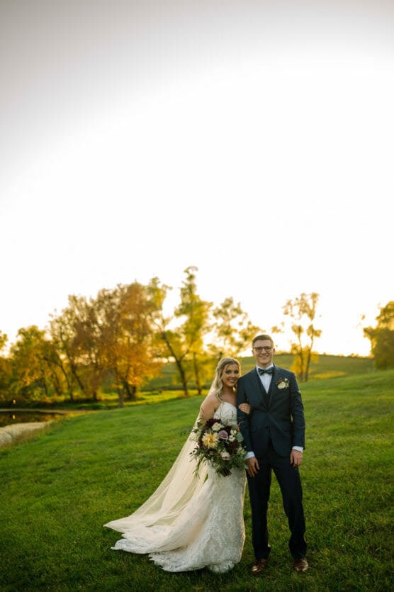 A bride in a white gown and a groom in a suit stand together on a grassy field at sunset, smiling and holding a bouquet. Autumn trees and a bright sky are in the background. by Justin Salem Meyer