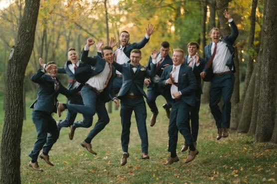 A group of groomsmen in suits jump joyfully in the air outdoors among trees, celebrating and smiling on a grassy area in a park during autumn. by Justin Salem Meyer