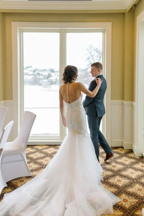 A bride in a white gown with a long train stands facing a groom in a suit, both near large windows with a snowy landscape outside. The bride gently holds the groom’s shoulder as they look at each other. by Justin Salem Meyer