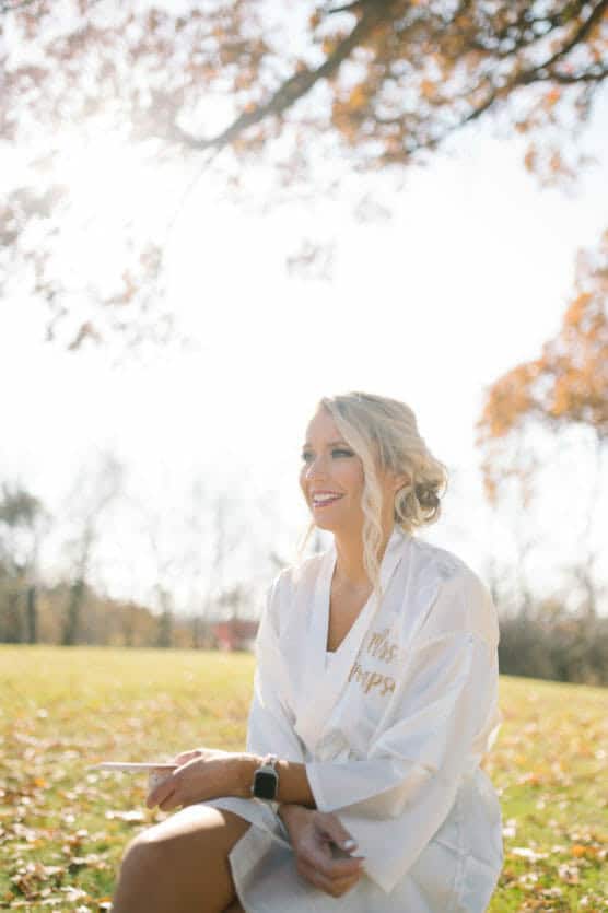 A smiling woman in a white satin robe with Mrs. embroidered on it sits outdoors on a sunny day, surrounded by autumn trees and fallen leaves, with sunlight filtering through the branches above. by Justin Salem Meyer