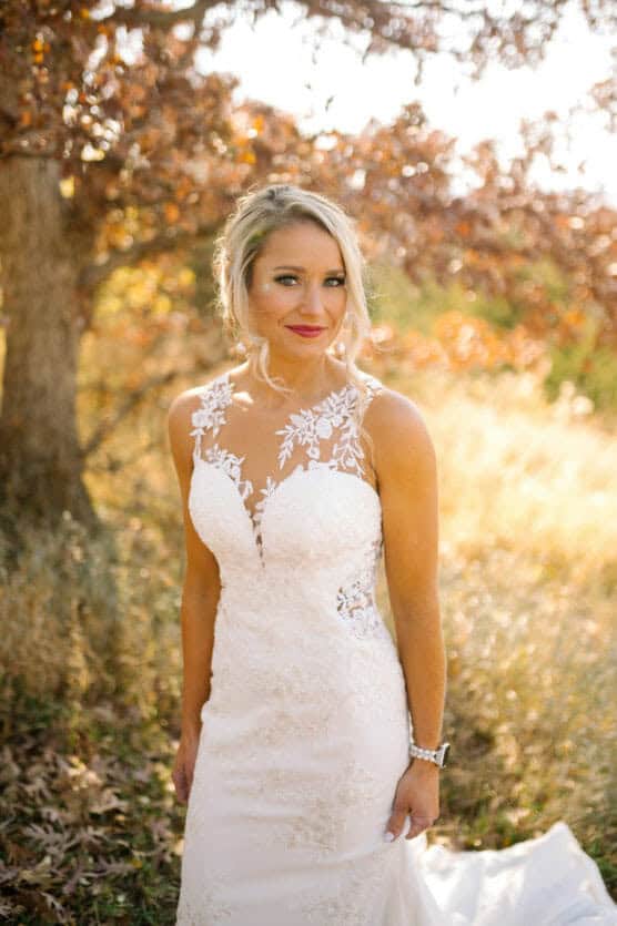 A woman in a white lace wedding dress stands outdoors in sunlight, surrounded by autumn trees and tall grass, smiling at the camera. by Justin Salem Meyer