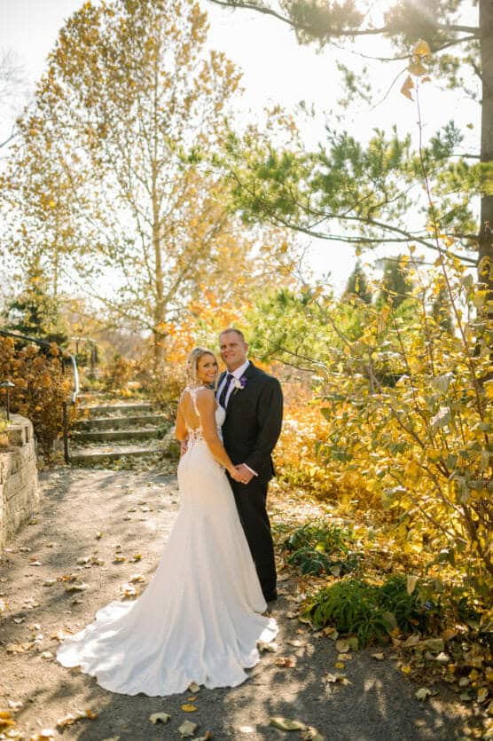 A bride in a white gown and a groom in a black suit stand close together on a sunlit garden path surrounded by autumn trees and golden foliage. by Justin Salem Meyer