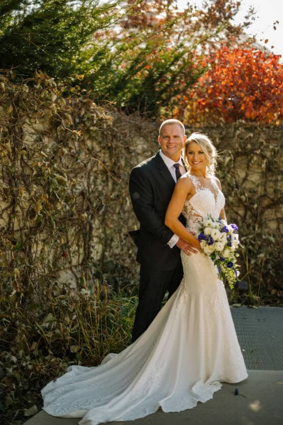 A bride in a white gown holding a bouquet stands smiling with a groom in a dark suit, both posing outdoors against a vine-covered wall with autumn foliage in the background. by Justin Salem Meyer