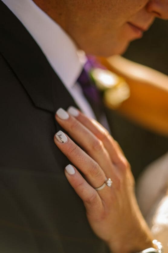 A close-up of a brides hand resting on a grooms suit jacket. She wears a diamond engagement ring and white nail polish; one nail has the word love written on it. The groom is dressed in a dark suit with a boutonnière. by Justin Salem Meyer