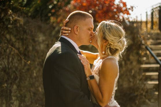 A bride and groom share a kiss outdoors in warm sunlight, surrounded by autumn foliage. The bride wears a white lace dress and the groom is in a dark suit. They embrace, standing close on their wedding day. by Justin Salem Meyer