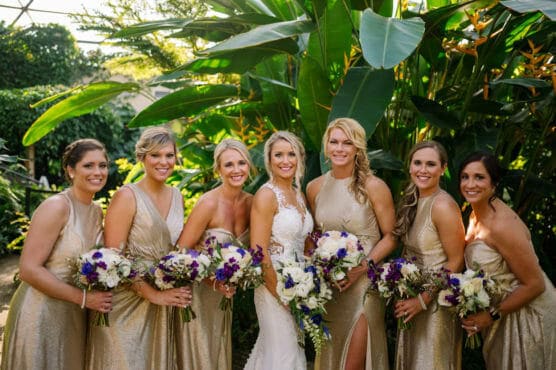 A bride in a white dress stands smiling with six bridesmaids in gold dresses, all holding bouquets of white and purple flowers, in front of lush green plants outdoors. by Justin Salem Meyer