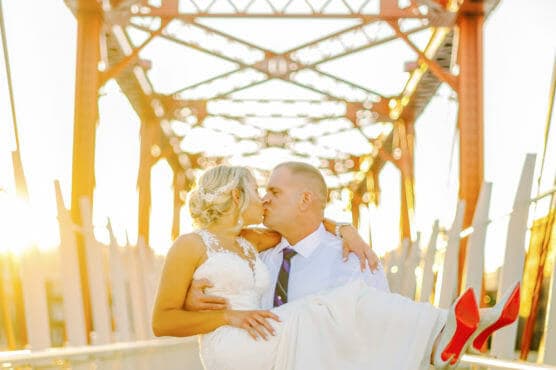 A groom in a white shirt and tie carries and kisses a bride in a white dress with red high heels on a sunlit, industrial-style bridge. The couple is framed by orange metal beams in the warm evening light. by Justin Salem Meyer