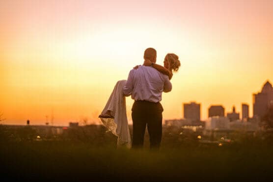 A man in formal attire carries a woman in a white dress while standing outdoors at sunset, with a city skyline in the background. by Justin Salem Meyer