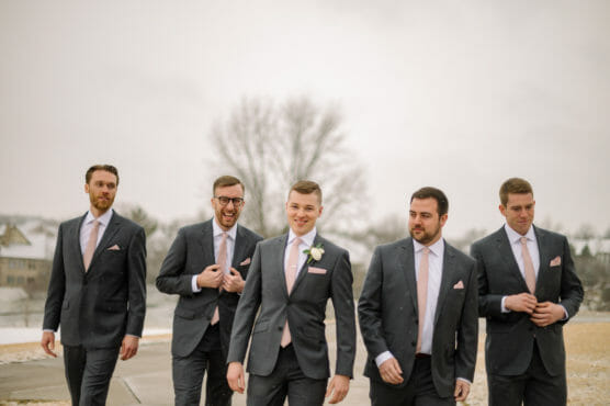 Five men in matching dark suits with pale pink ties and pocket squares walk outside on a cloudy day, smiling and laughing. Snow covers the ground, and bare trees can be seen in the background. by Justin Salem Meyer