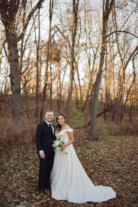 A bride in a white off-the-shoulder gown and a groom in a dark suit stand together in a forest with bare trees and fallen leaves, smiling at the camera. by Justin Salem Meyer