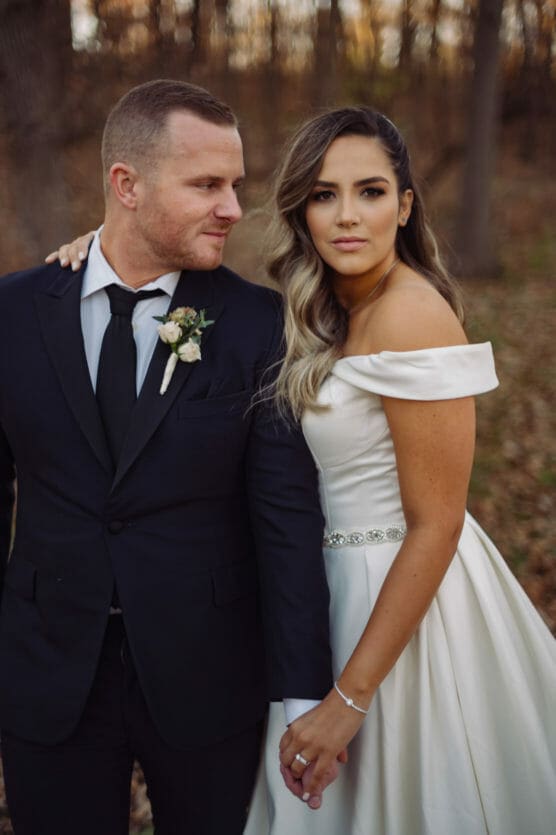 A bride in an off-shoulder white gown and a groom in a black suit hold hands outdoors in a wooded area, looking in different directions with serious expressions. by Justin Salem Meyer