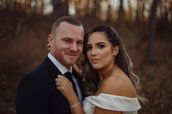 A couple poses closely outdoors, both looking at the camera. The woman wears an off-shoulder white dress and jewelry, while the man wears a dark suit and tie. The background is blurred with trees and autumn colors. by Justin Salem Meyer
