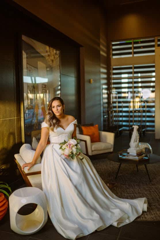 A bride in an off-the-shoulder white gown holds a bouquet of flowers while sitting on a modern chair in a stylish, warmly lit lounge with contemporary decor. by Justin Salem Meyer