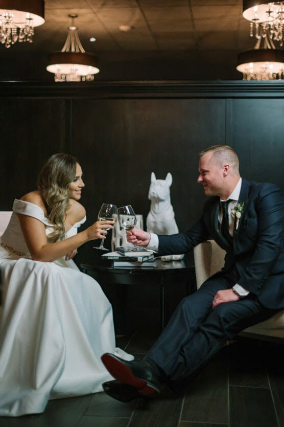 A bride and groom in formal attire sit across from each other, smiling and toasting with wine glasses. They are indoors with elegant lighting, and a white dog statue is visible on the table between them. by Justin Salem Meyer
