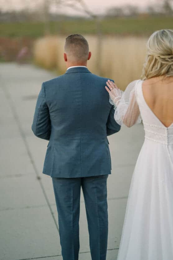A bride in a white dress gently touches the shoulder of a groom in a blue suit from behind, outdoors on a paved walkway. by Justin Salem Meyer