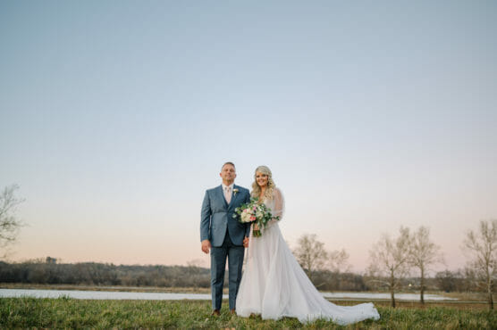 A bride in a long white dress holding a bouquet stands next to a man in a blue suit outdoors on grass, with a calm river, bare trees, and a clear sky at sunset in the background. by Justin Salem Meyer