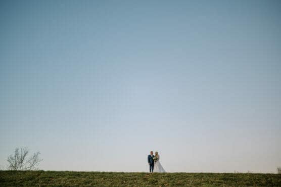 A bride and groom stand close together on a grassy hill under a vast, clear blue sky, with minimal landscape and no other people or objects around them. by Justin Salem Meyer